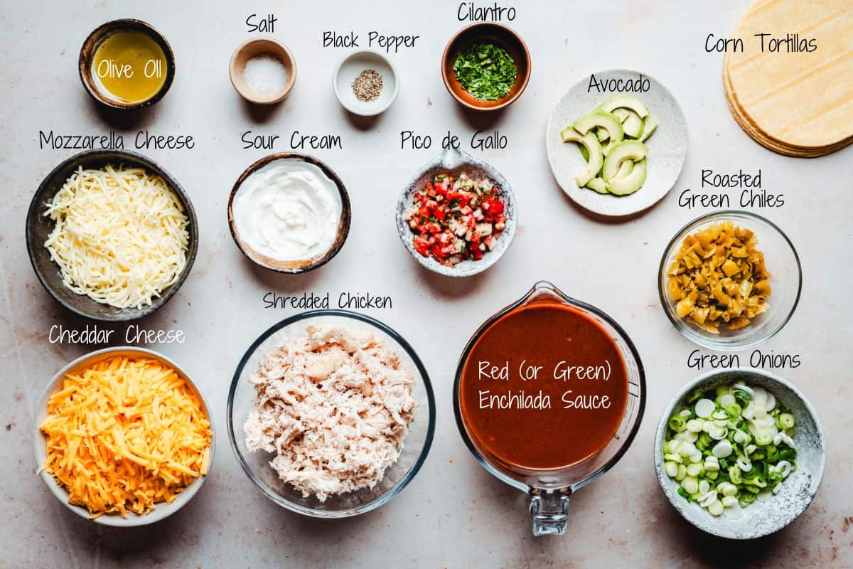 An assortment of ingredients is laid out for making slow cooker chicken enchiladas, including olive oil, salt, black pepper, cilantro, corn tortillas, mozzarella and cheddar cheese, sour cream, pico de gallo, shredded chicken, enchilada sauce, avocado, green onions, and roasted green chiles.