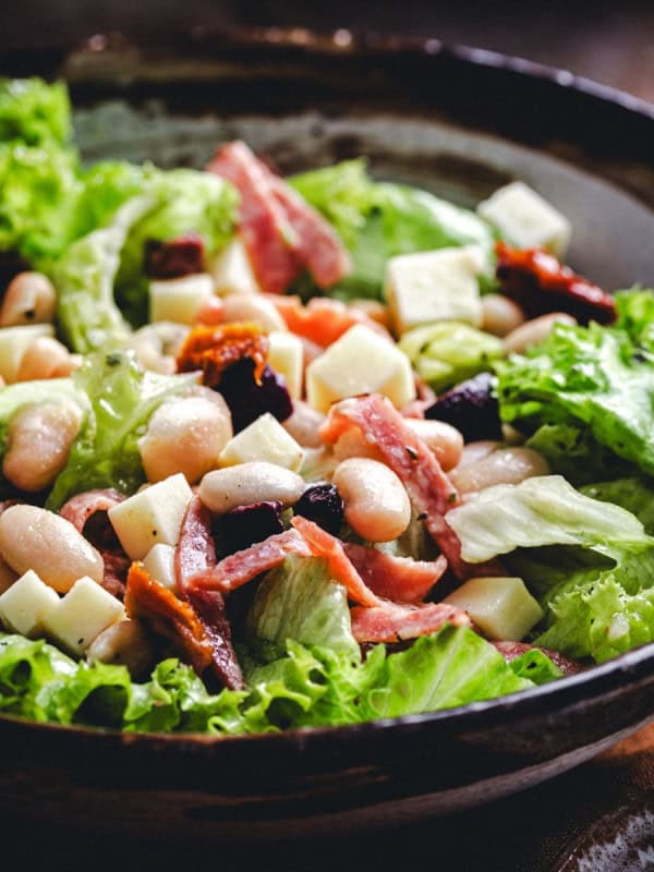 A bowl of fresh salad with large green lettuce leaves, white beans, chunks of cheese, pieces of ham or bacon, and sun-dried tomatoes, served in a rustic dark bowl. The ingredients are evenly mixed, creating a colorful and appetizing dish.