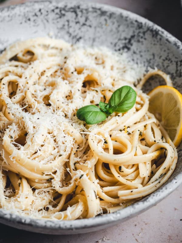 A bowl of creamy spaghetti topped with grated cheese, black pepper, and garnished with a fresh basil leaf and a lemon slice. The dish is served in a speckled, rustic bowl with a fork resting inside.