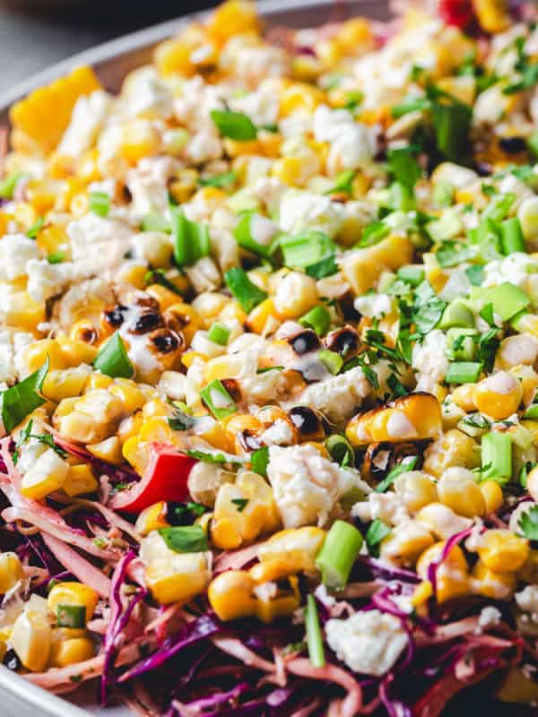 A colorful Mexican Street Corn Salad on a plate, featuring charred corn, crumbled feta cheese, chopped green onions, and red cabbage. The salad is garnished with fresh herbs, and drizzled with a dark balsamic glaze or dressing. The vibrant mix showcases a variety of textures and colors.