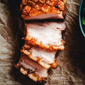 A close-up of sliced, juicy pork belly with crispy, golden-brown skin, arranged on a piece of parchment paper. The tender, perfectly cooked layers of meat and fat are clearly visible, with a portion of fresh greens on a plate partially seen on the right.