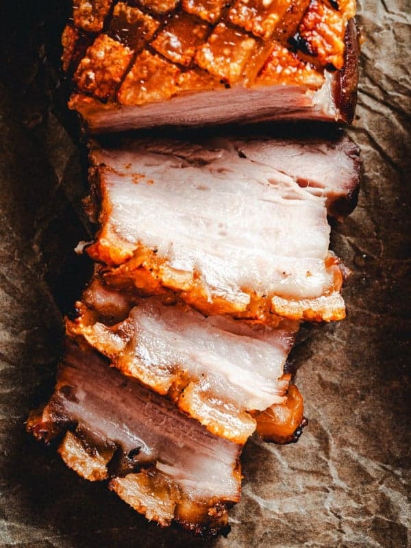 A close-up of sliced, juicy pork belly with crispy, golden-brown skin, arranged on a piece of parchment paper. The tender, perfectly cooked layers of meat and fat are clearly visible, with a portion of fresh greens on a plate partially seen on the right.