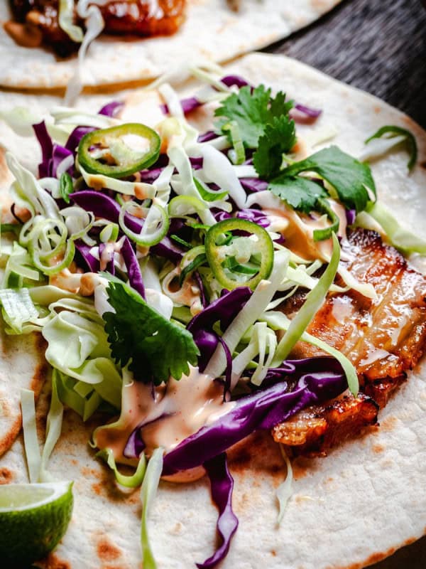 Close-up of a taco with grilled pork belly strips, shredded green and purple cabbage, sliced jalapeños, fresh cilantro, and a drizzle of creamy sauce on a flour tortilla. A lime wedge is partially visible to the side.