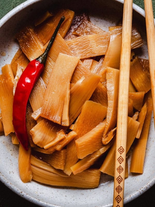 A ceramic bowl filled with marinated bamboo shoots, topped with a whole red chili pepper. Wooden chopsticks are placed on the rim of the bowl against a dark, textured background.