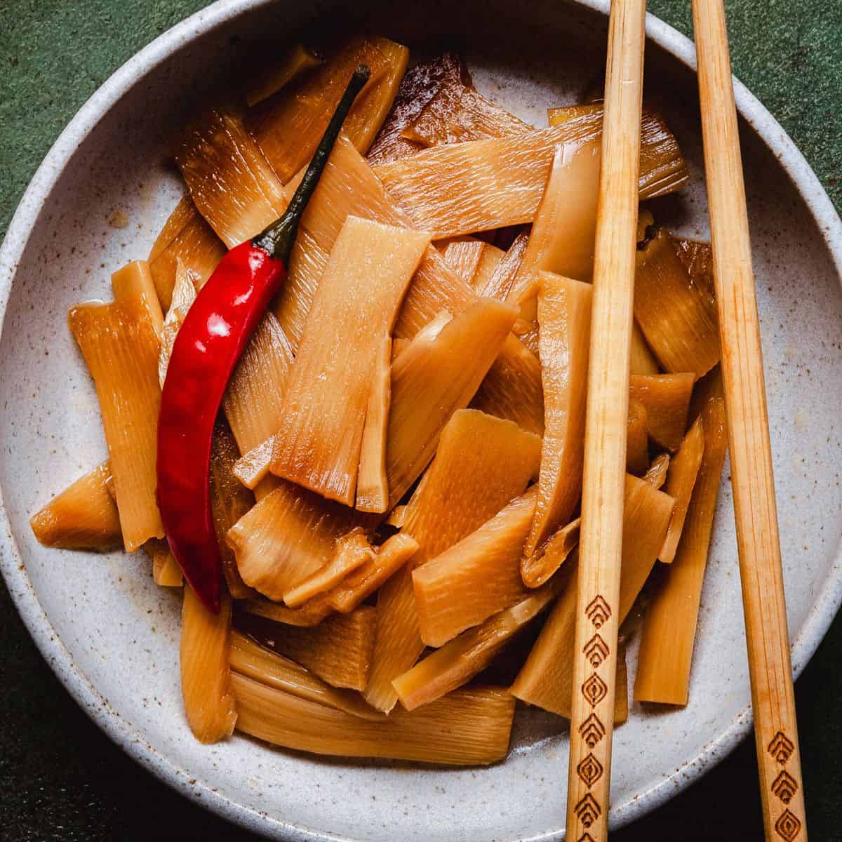 A ceramic bowl filled with marinated bamboo shoots, topped with a whole red chili pepper. Wooden chopsticks are placed on the rim of the bowl against a dark, textured background.