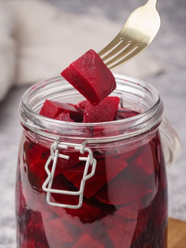 A fork holding up a cube of pickled beets above an open glass jar filled with more beet cubes. The jar has a metal clasp and sits on a gray surface, with a white cloth blurred in the background.