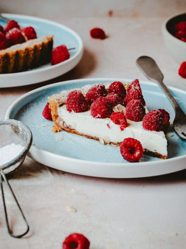 A slice of raspberry tart with a creamy filling on a blue plate, garnished with fresh raspberries and powdered sugar. A metal fork rests on the plate. Additional tart slices, raspberries, a strainer with powdered sugar, and another plate are visible in the background.