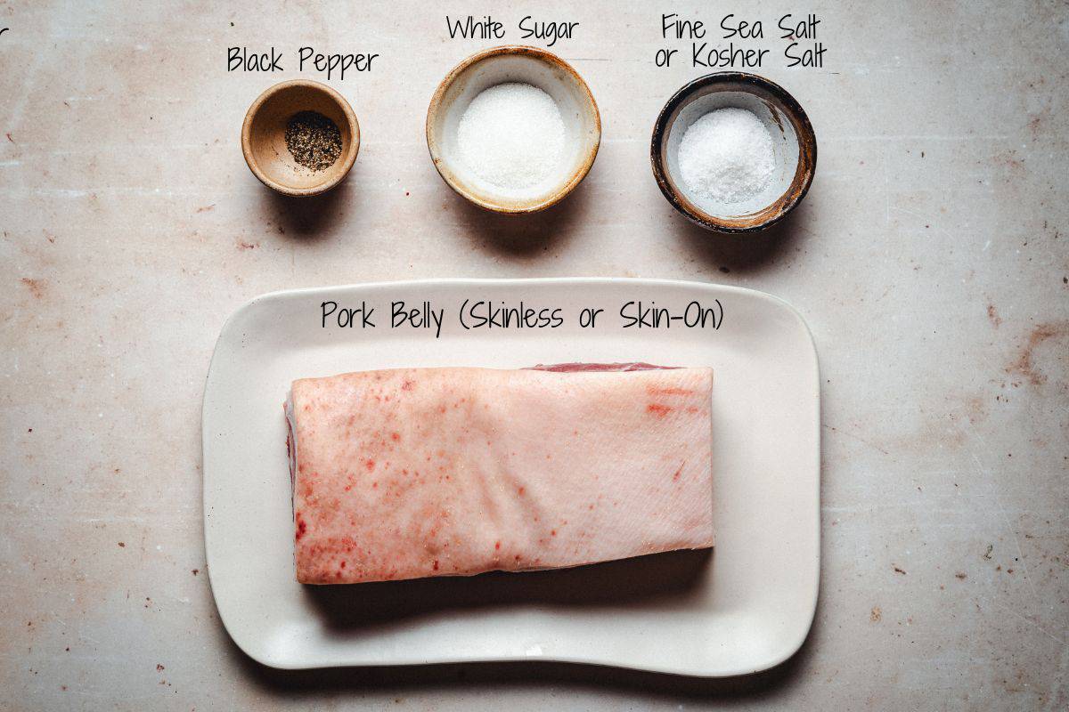 Image showing a raw piece of pork belly on a plate. Above it are three small bowls containing black pepper, white sugar, and fine sea salt or kosher salt. Each bowl is labeled with black handwritten text identifying the contents. The background is a light-colored surface.