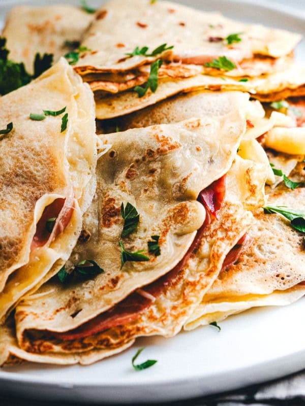 A close-up of a plate stacked with savory crepes, folded and filled with what appears to be ham and cheese. The crepes are garnished with fresh parsley leaves, adding a touch of color to the dish.