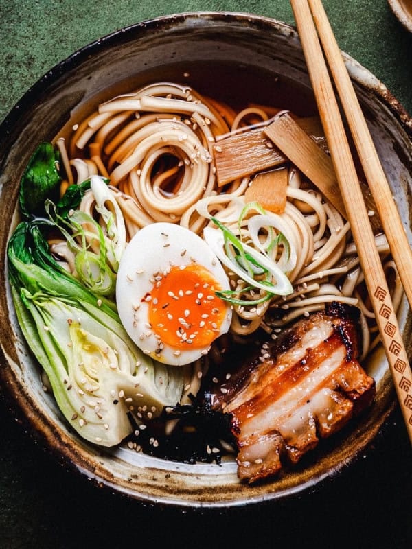 A bowl of ramen with noodles, bok choy, a soft-boiled egg, pork belly, bamboo shoots, and green onions, garnished with sesame seeds. Chopsticks rest on the bowl, and small dishes are in the background.