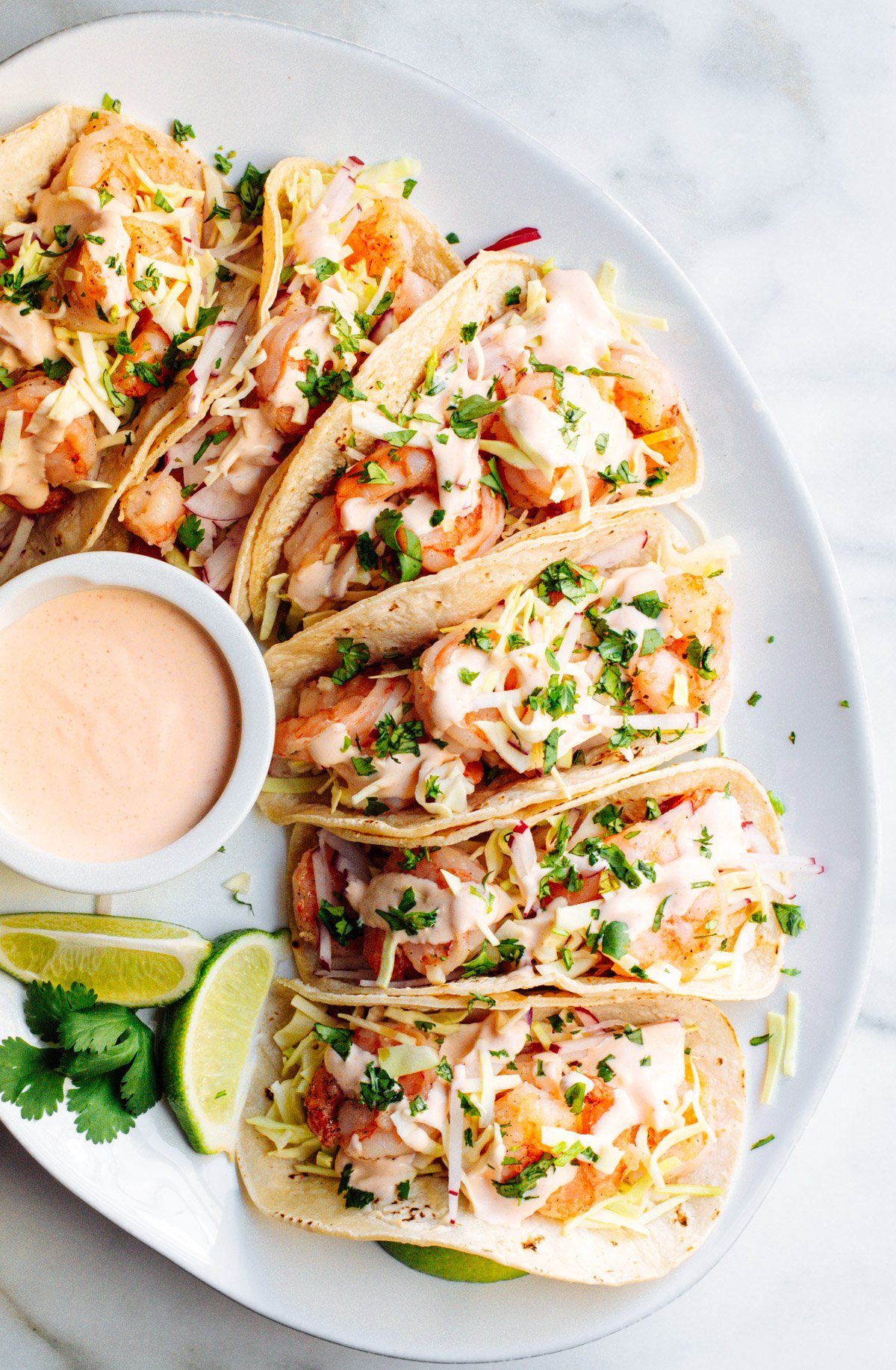 Shrimp Tacos served on a white oval platter with extra sauce and lime wedges on the side.