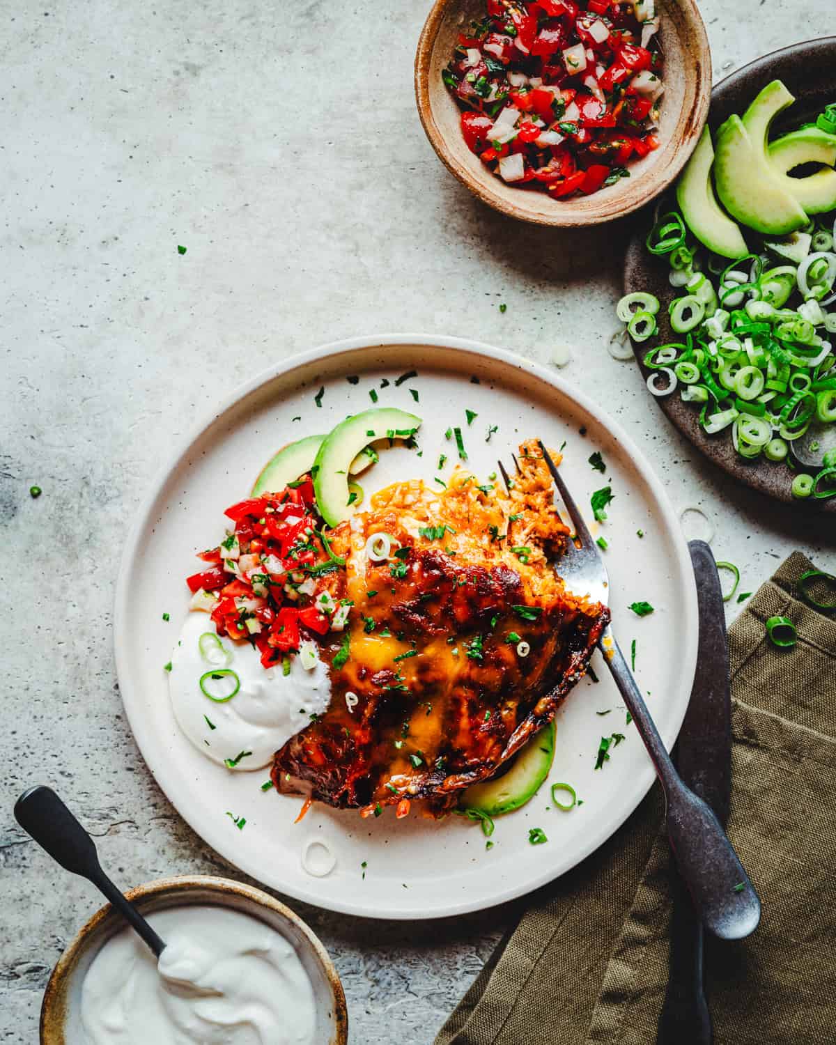 A plate of enchiladas topped with melted cheese, chopped green onions, and pico de gallo, garnished with avocado slices and a dollop of sour cream on the side. Bowls of salsa, sliced avocados, and sour cream are placed nearby.