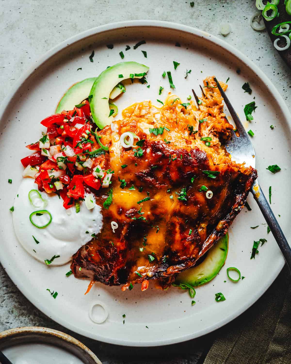 A plate featuring a serving of cheesy enchiladas garnished with chopped cilantro and green onions. Slices of avocado and a scoop of sour cream are on the side, accompanied by a colorful pico de gallo mix of diced tomatoes and onions.