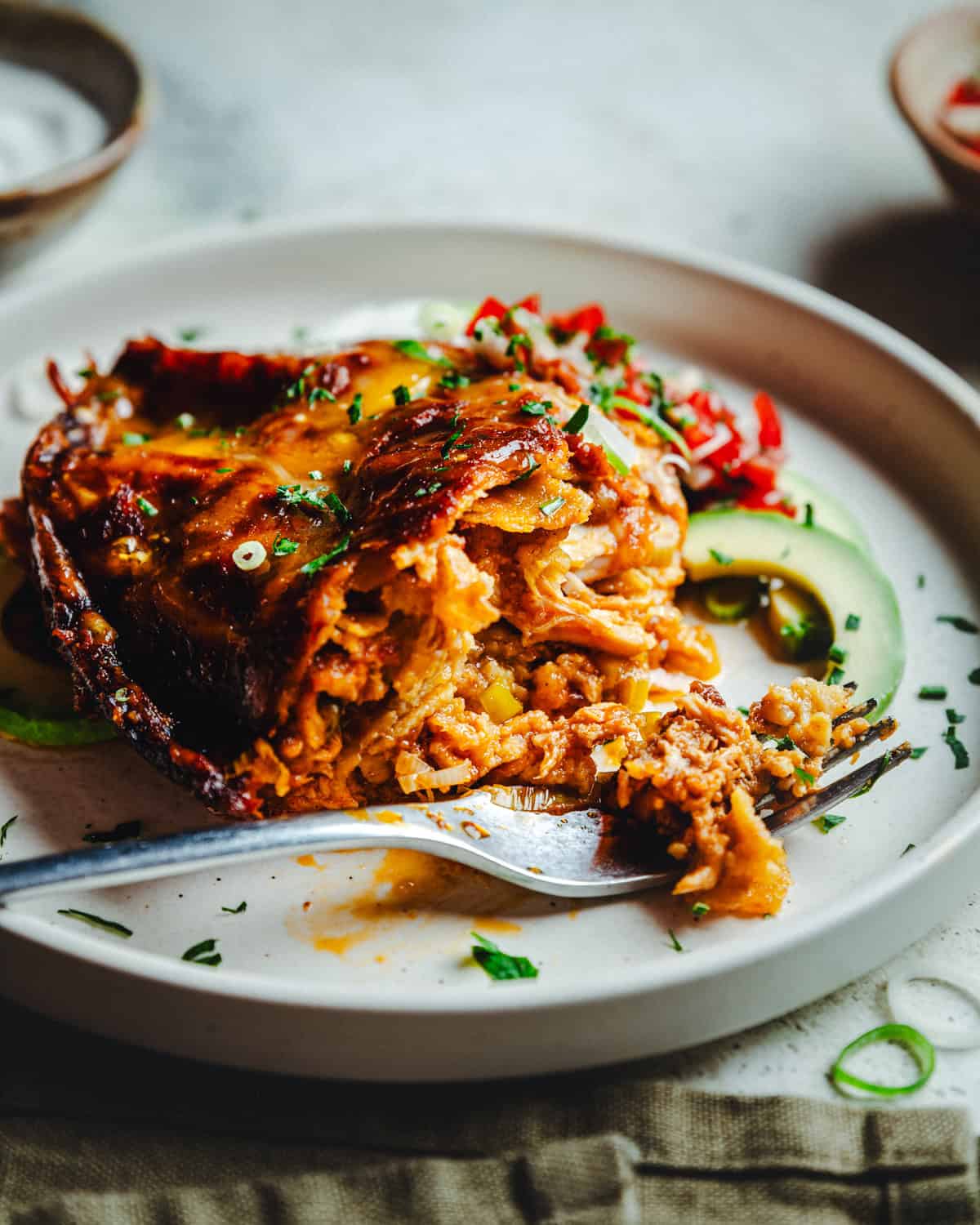 Enchiladas, topped with melted cheese, is served on a white plate. Freshly sliced cucumbers and chopped herbs garnish the dish. A fork rests on the plate, ready for a bite.