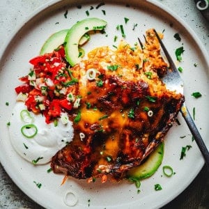 A plate with a serving of chicken enchiladas covered in melted cheese, garnished with sliced green onions and parsley. Accompanied by sliced avocado, dollops of sour cream, and a portion of diced tomatoes and green herbs. A fork rests on the plate.