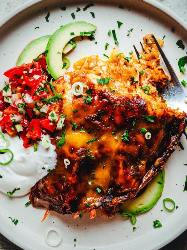 A plate with a serving of chicken enchiladas covered in melted cheese, garnished with sliced green onions and parsley. Accompanied by sliced avocado, dollops of sour cream, and a portion of diced tomatoes and green herbs. A fork rests on the plate.