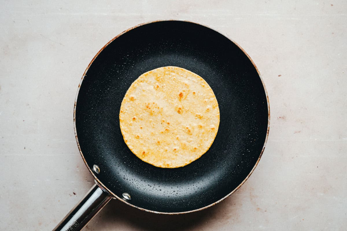 A single corn tortilla is heating in a black non-stick frying pan on a light-colored surface.