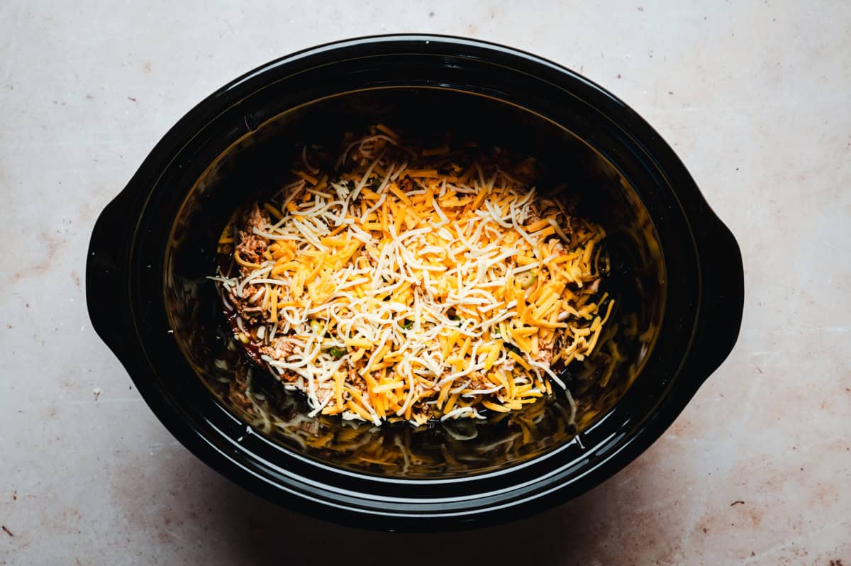 A slow cooker filled with shredded cheese, layered over other food ingredients. The cheese is a mix of yellow and white varieties, spread evenly on top. The slow cooker is set on a light-colored countertop.