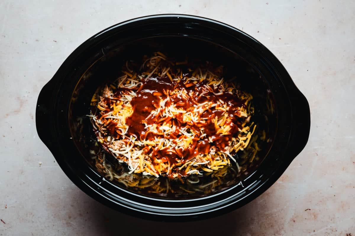 A black slow cooker filled with shredded cheese and a rich red enchilada sauce, ready to cook. The ingredients are spread evenly inside the pot, creating a colorful and appetizing mixture.