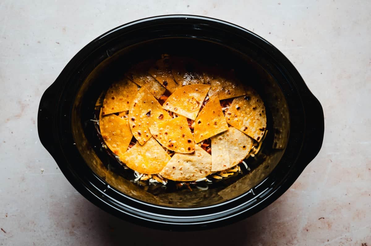 A black slow cooker filled with layered, seasoned corn tortillas on top of enchilada filling. The tortillas are golden brown, slightly crispy.