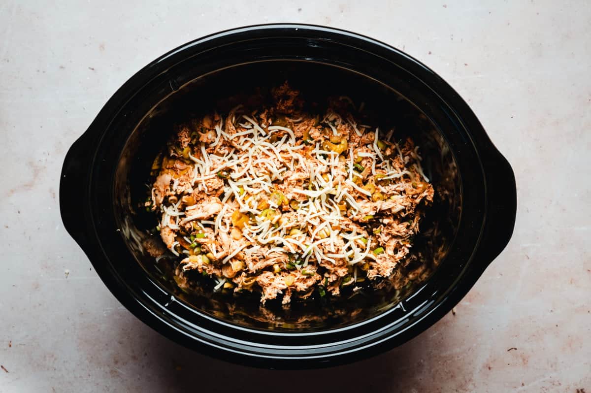 A black slow cooker filled with enchilada filling and topped with cheese, resting on a light-colored countertop.