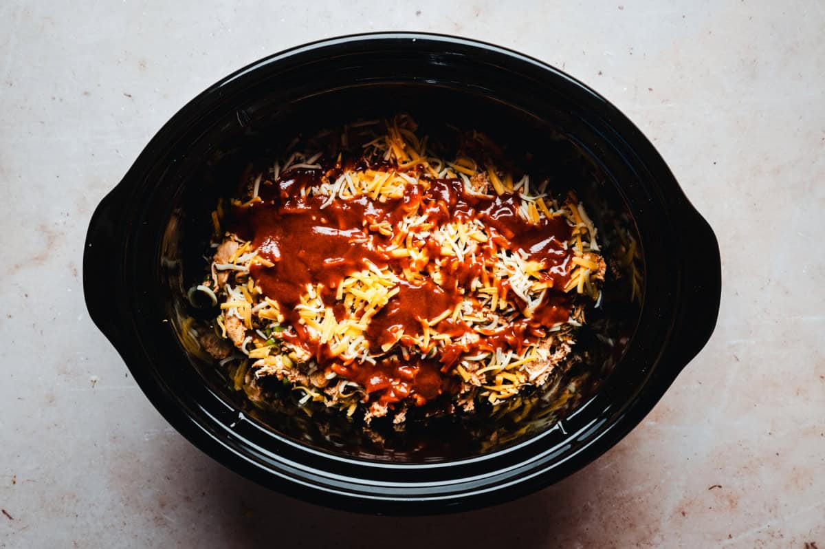 A slow cooker filled with enchilada filling topped with sauce and melted cheese. The dish sits on a light-colored countertop, showcasing a rich blend of colors and textures.