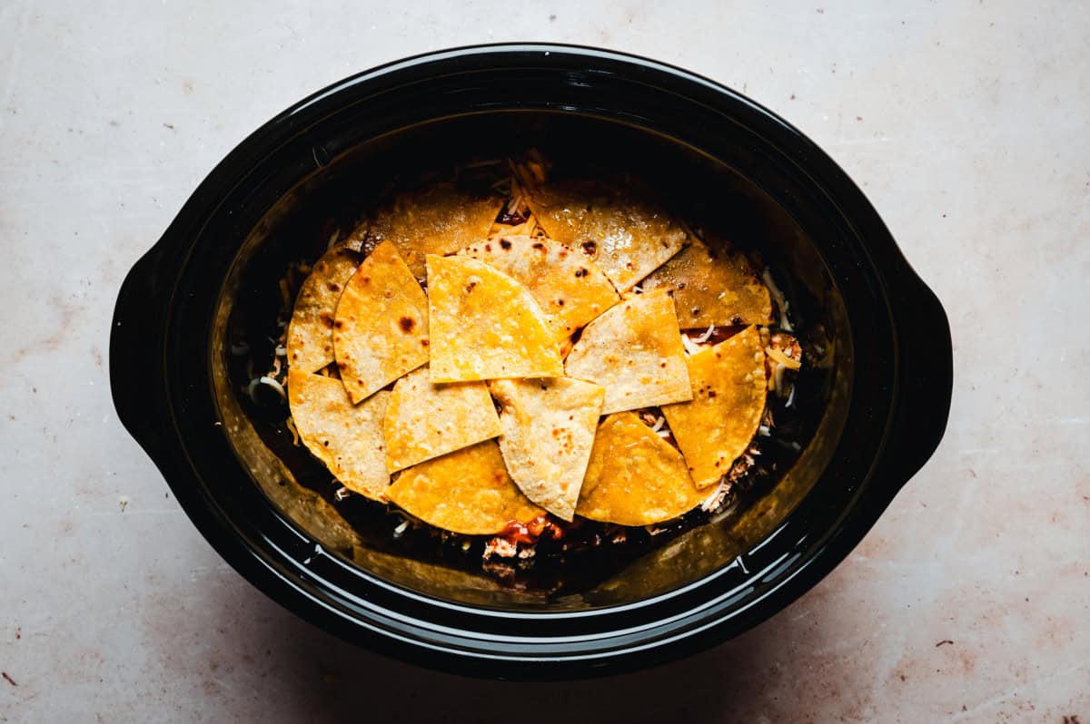 A slow cooker filled with layered ingredients, topped with triangular tortilla chips.