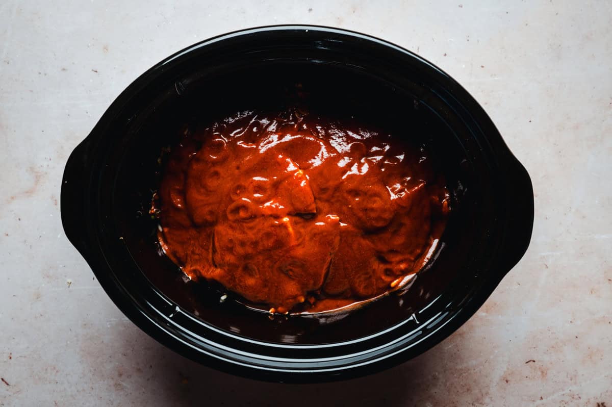 A black slow cooker filled layered ingredients covered in a rich, red enchilada sauce, seen from a top-down angle on a light countertop.