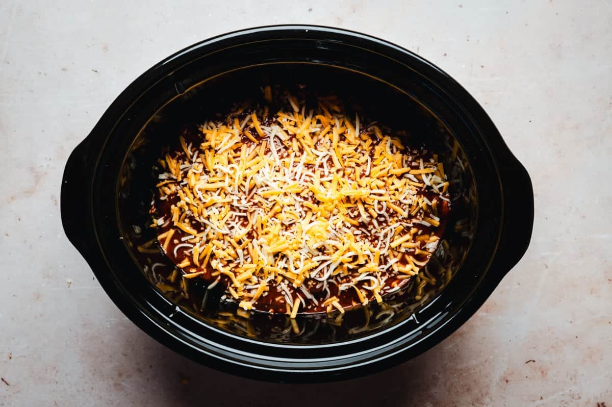 A black slow cooker filled with a dish topped with melted cheddar cheese, set on a light-colored countertop.