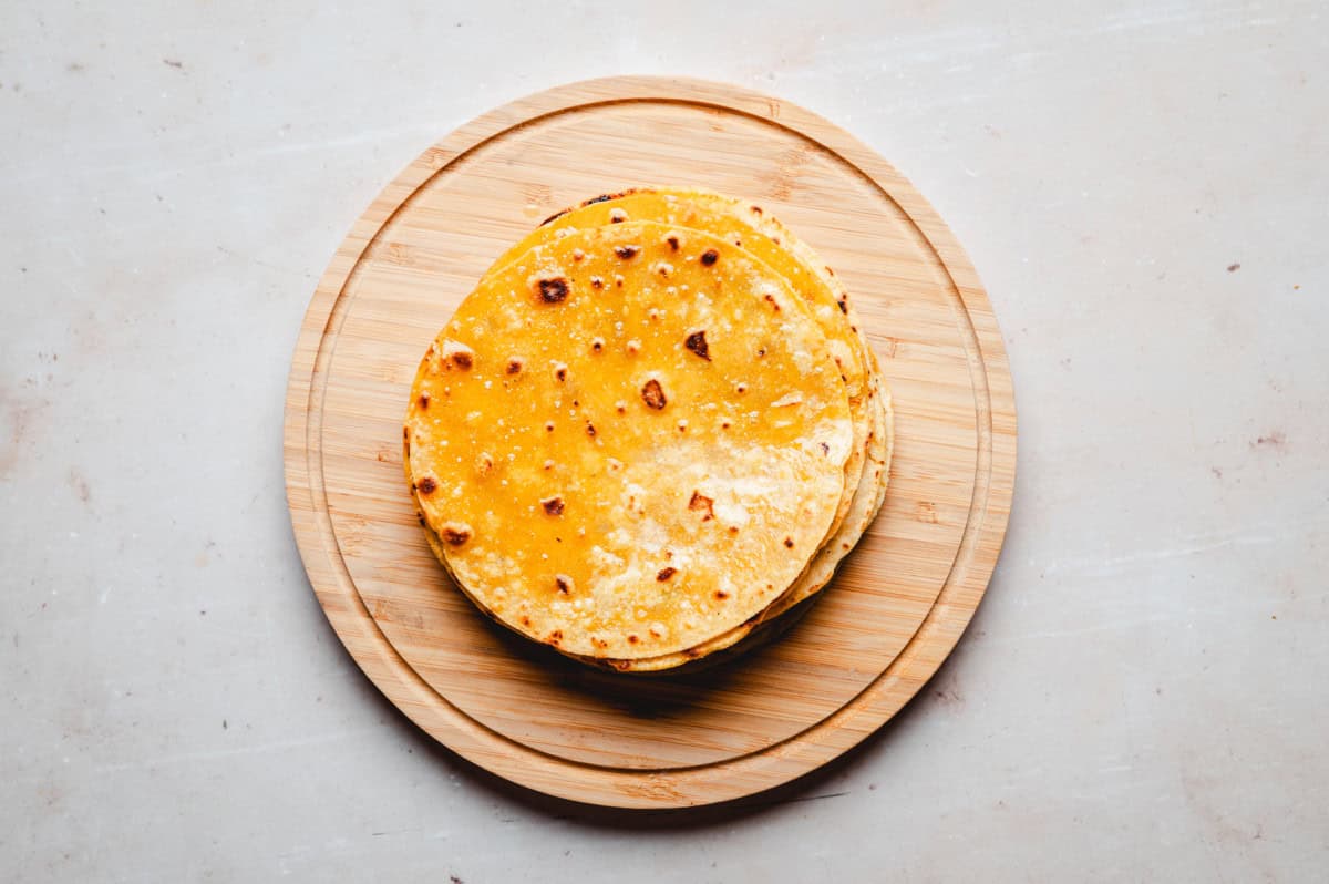 A stack of golden corn tortillas on a round wooden board against a light background.