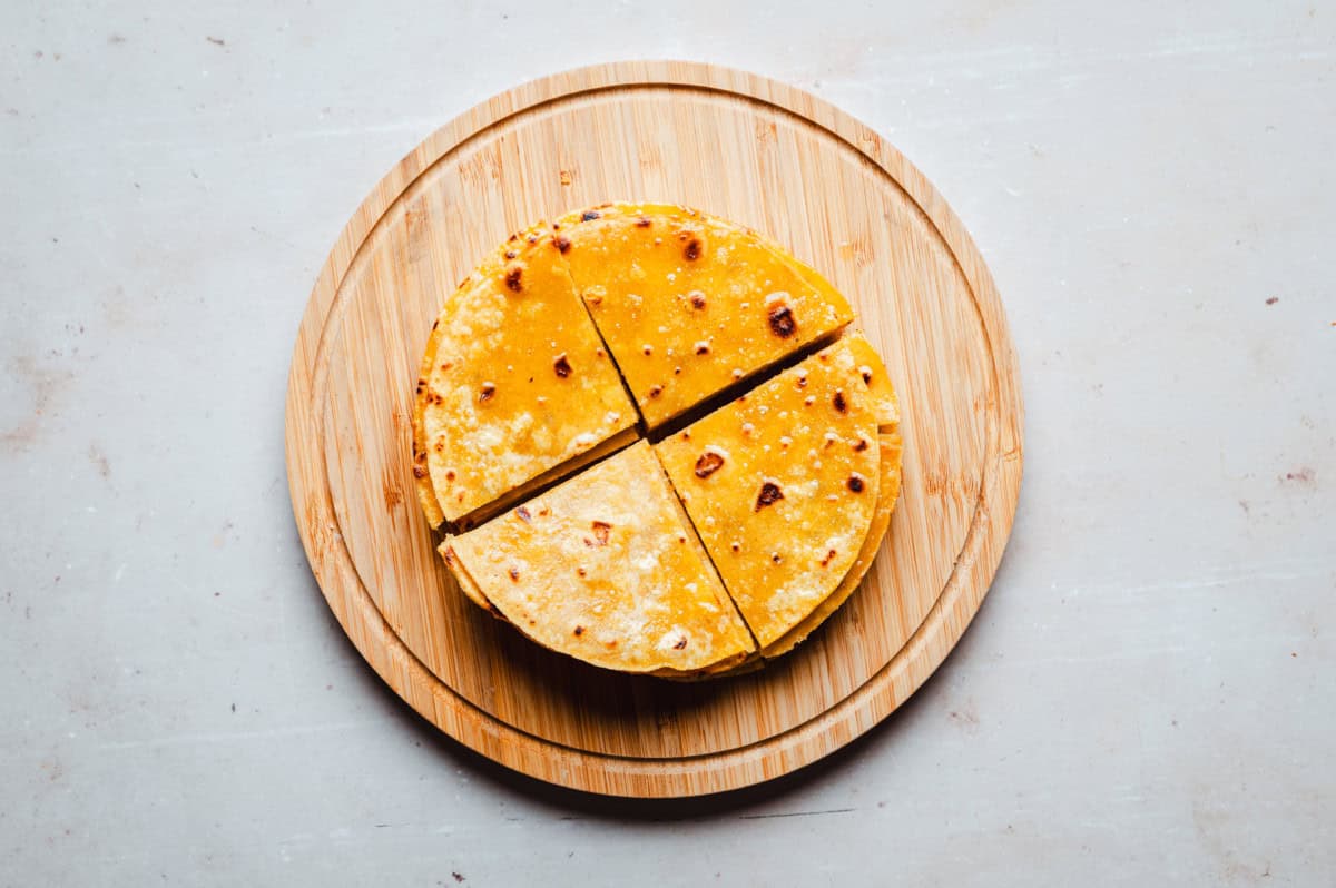 A stack of tortillas, neatly cut into quarters, is placed on a round wooden board. The tortillas have a golden brown color and a few scattered brown spots, suggesting they are freshly cooked. The board rests on a light-colored surface.