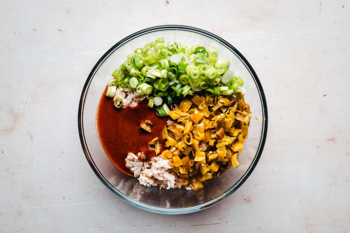 A glass bowl containing minced chicken, red enchilada sauce, chopped green onions, and diced green peppers on a light-colored surface.