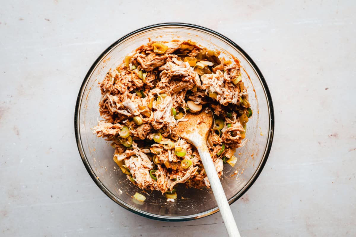 A glass bowl filled with a mixture of pulled chicken, enchilada sauce and green chiles. A wooden spoon rests inside the bowl, stirring the ingredients together on a light-colored surface.