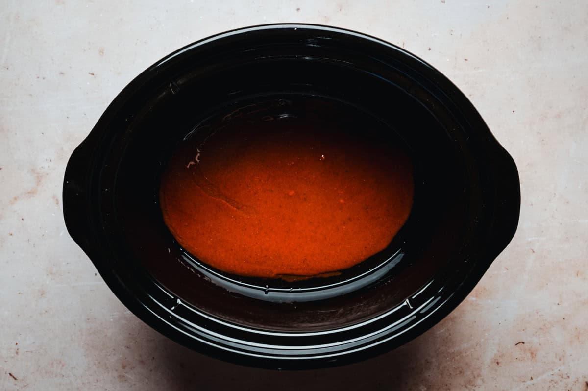 A slow cooker with a glossy, deep red enchilada sauce inside. The sauce is smooth and evenly distributed, and the slow cooker sits on a light, textured surface.