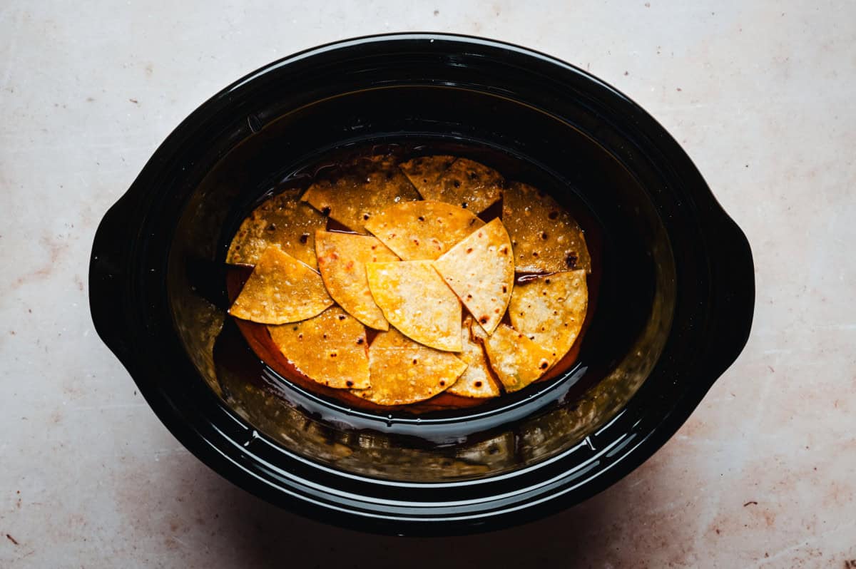 A black slow cooker filled with circular slices of tortillas layered on top of enchilada sauce. The tortillas have a golden, crispy appearance, and the cooker is set against a light, textured background.