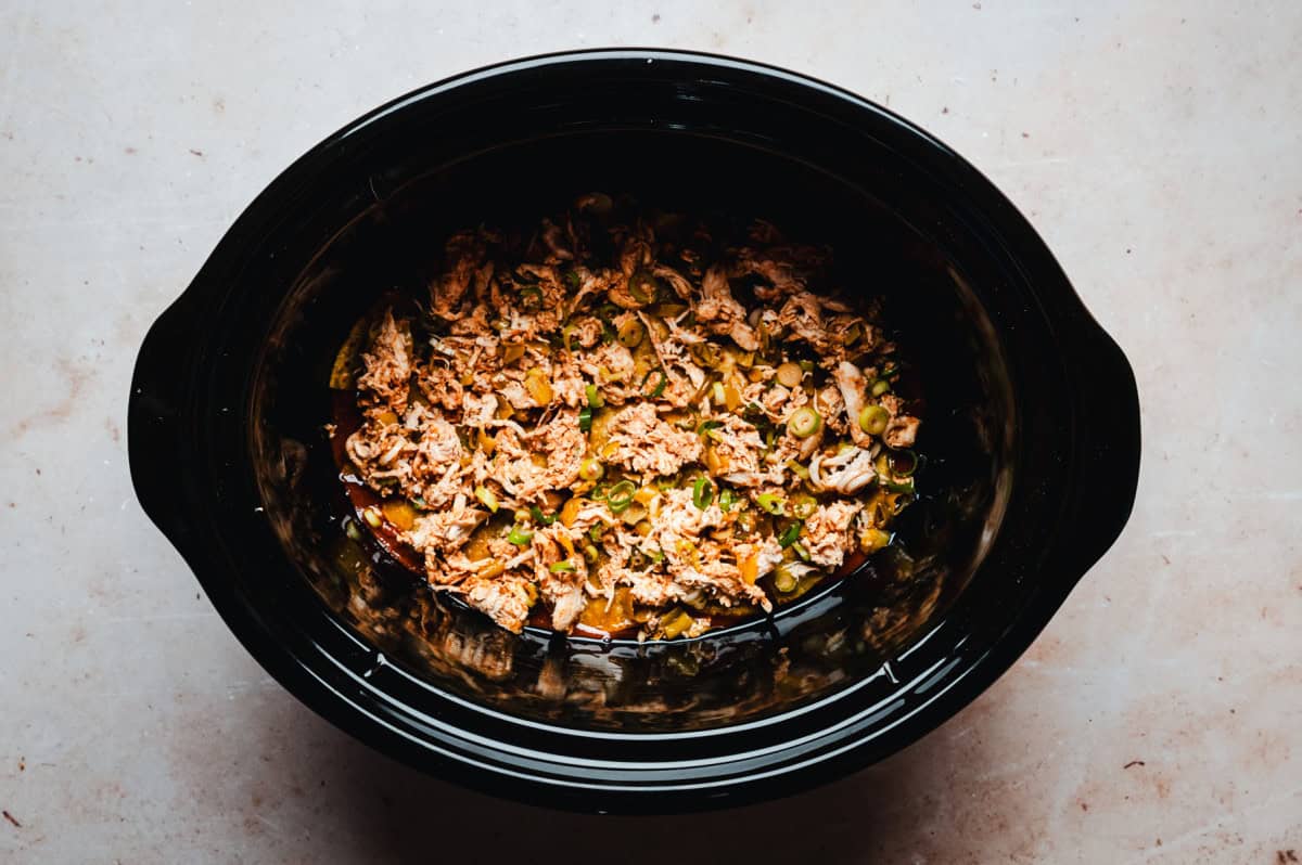 Top view of a dark slow cooker filled with shredded meat and enchilada ingredients. The cooker rests on a light marble surface.