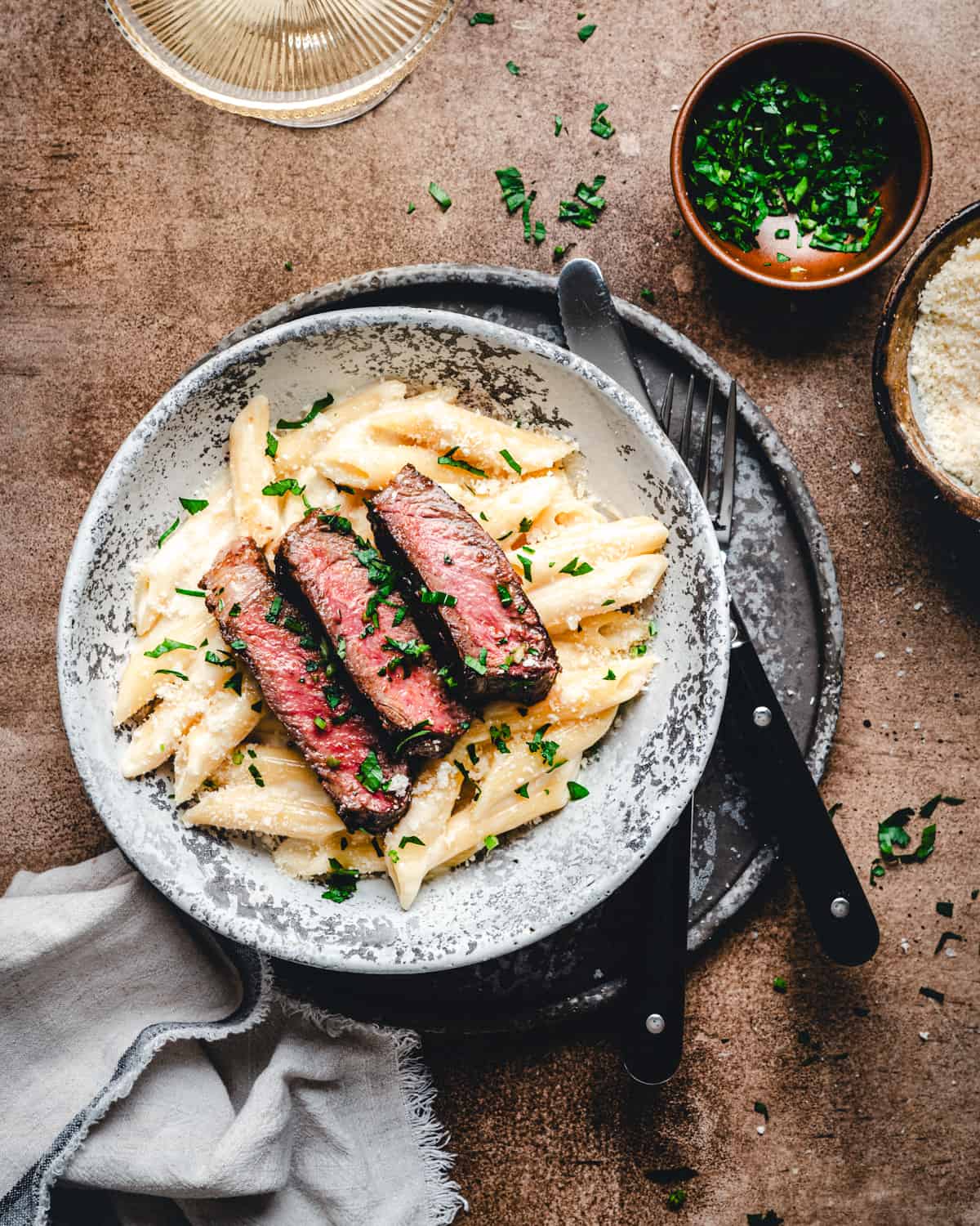 Slices of seared ribeye steak over creamy penne pasta in a light gray bowl photographed from the top.