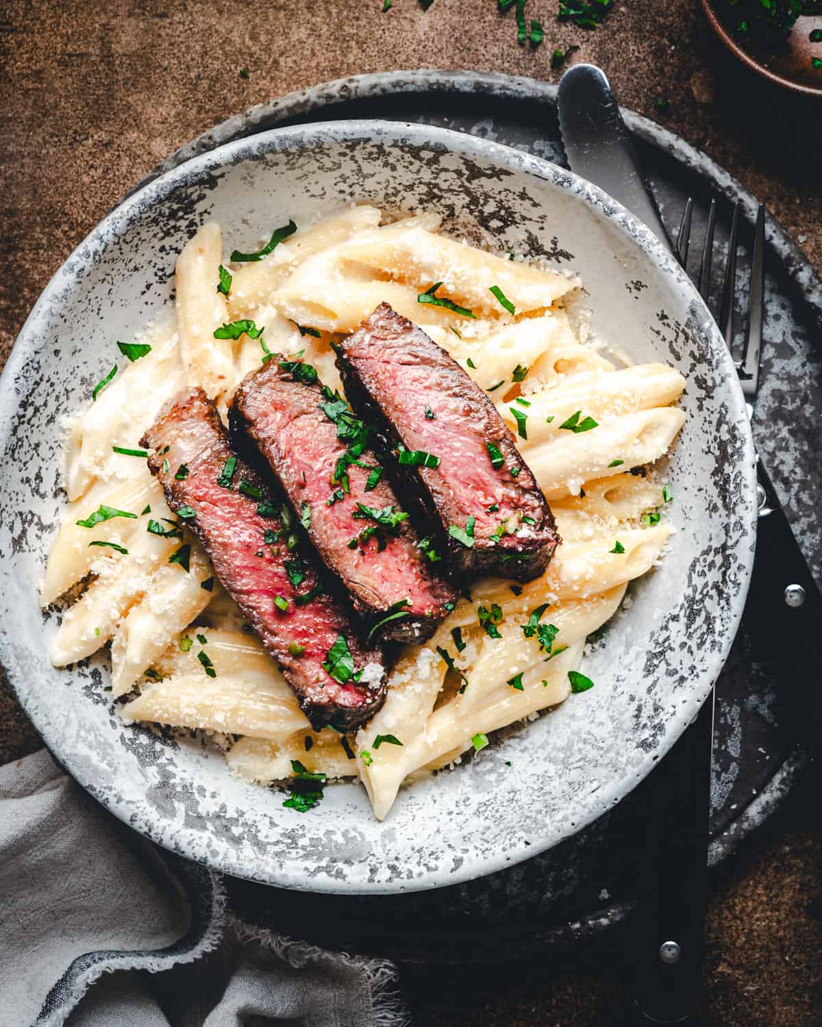 Slices of seared ribeye steak over creamy penne pasta in a light gray bowl photographed from the top close up.