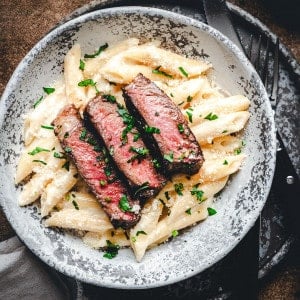 Slices of seared ribeye steak over creamy penne pasta in a light gray bowl photographed from the top close up.