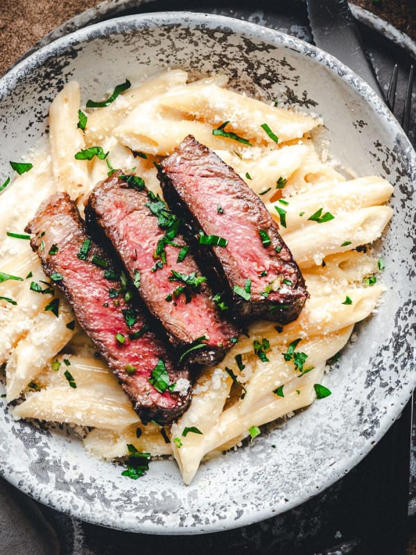 Slices of seared ribeye steak over creamy penne pasta in a light gray bowl photographed from the top close up.