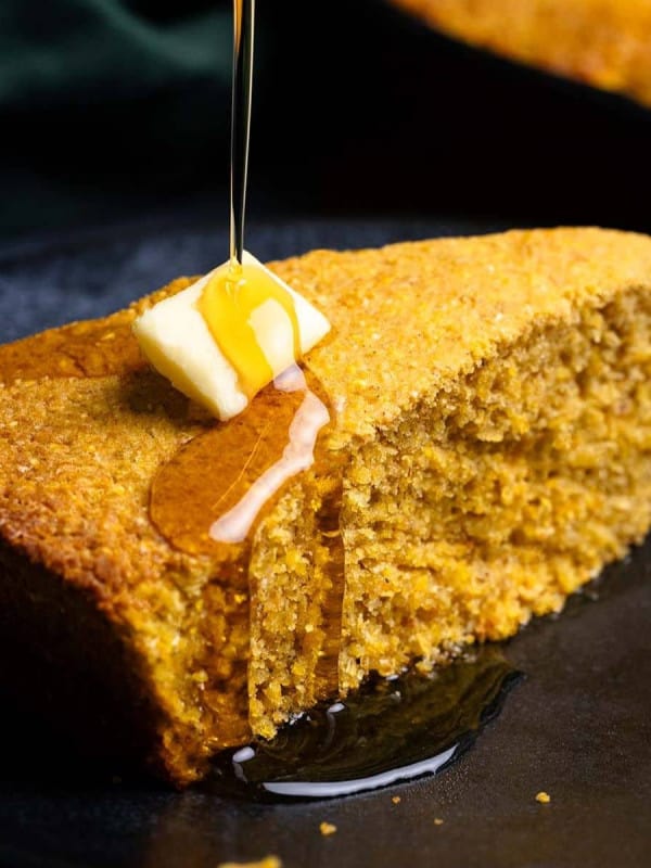 A slice of Sweet Potato Cornbread on a black plate with a pat of butter and drizzle of maple syrup.