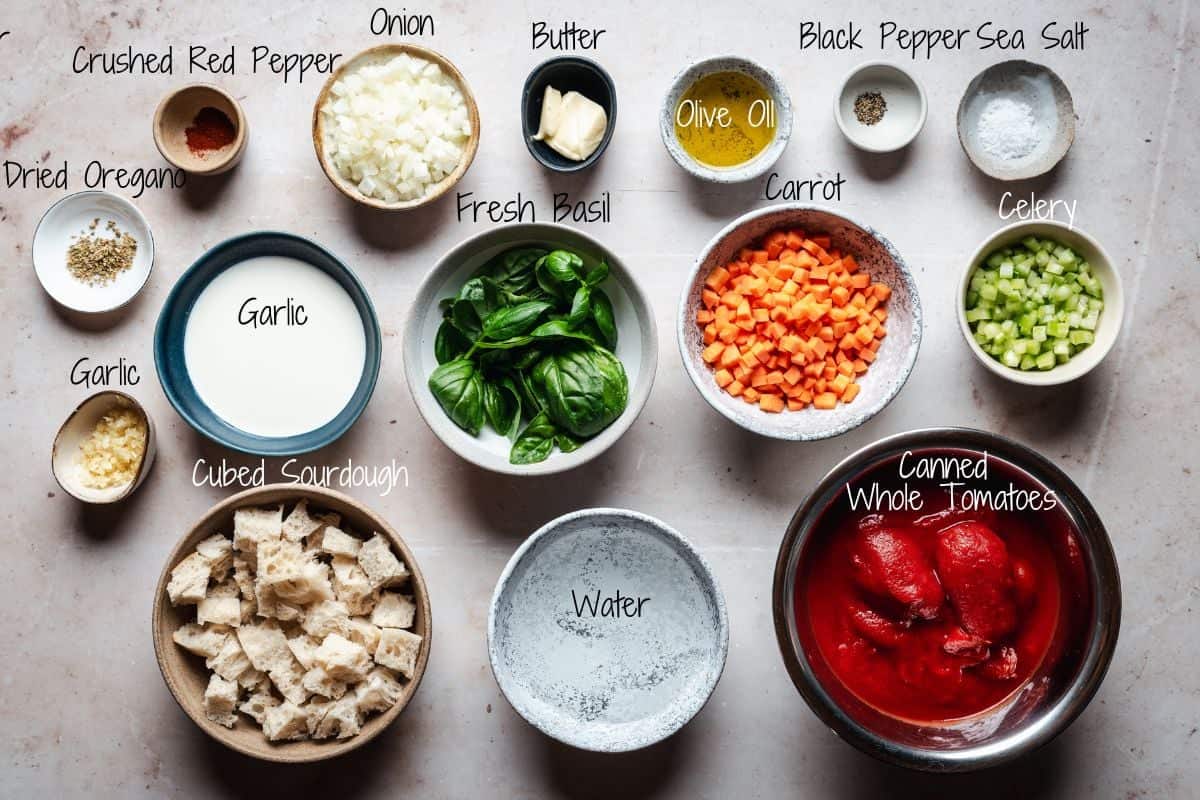 Ingredients for a recipe are arranged on a table, including canned whole tomatoes, cubed sourdough, water, cubed carrots, celery, chopped onion, minced garlic, crushed red pepper, dried oregano, fresh basil, pepper, salt, olive oil, and butter.