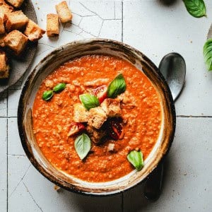 A bowl of tomato soup garnished with croutons, fresh basil leaves, and cherry tomato slices. A black spoon lies beside the bowl on a tile surface, with a small plate of croutons to the side.