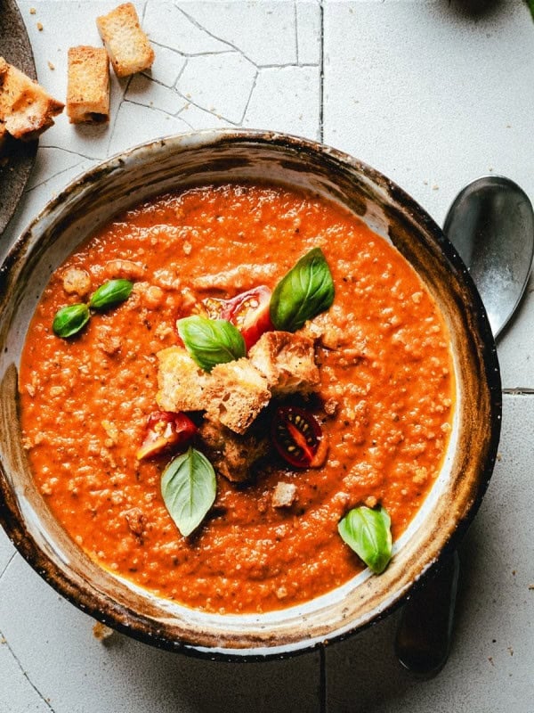 A bowl of tomato soup garnished with croutons, fresh basil leaves, and cherry tomato slices. A black spoon lies beside the bowl on a tile surface, with a small plate of croutons to the side.