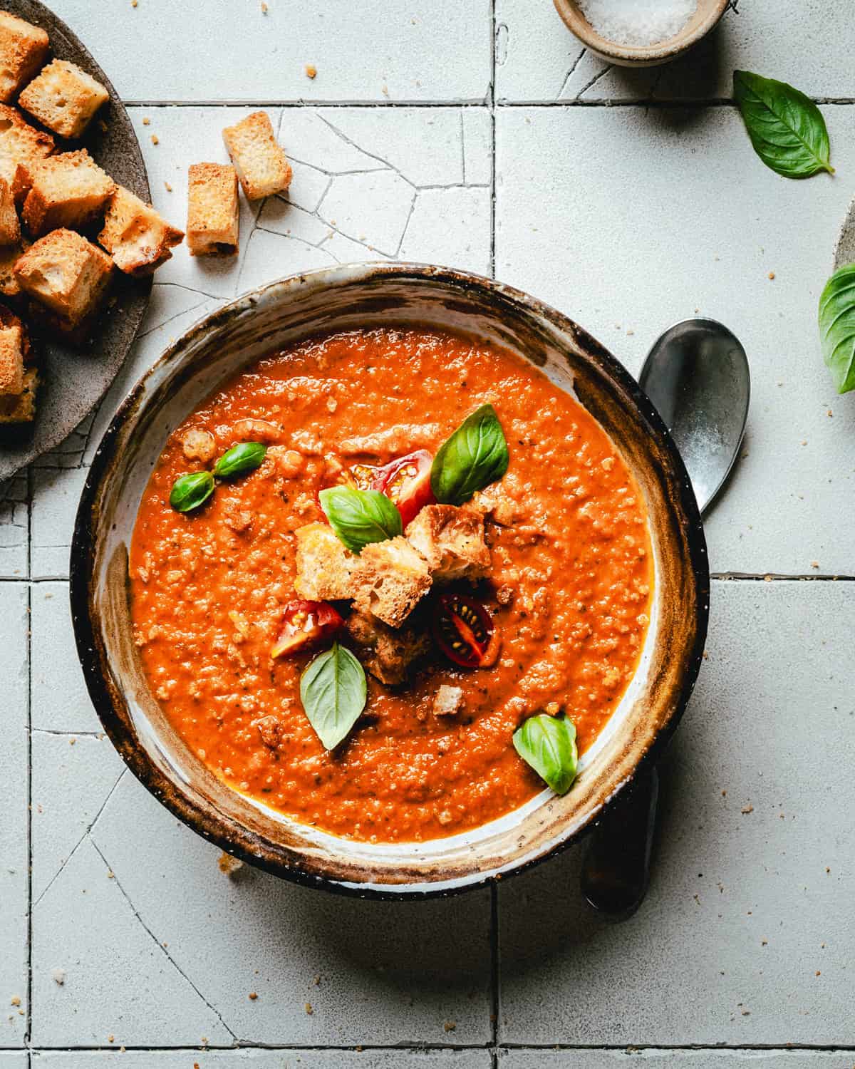 A rustic bowl of tomato soup garnished with croutons, sliced tomato, and fresh basil leaves sits on a cracked white tile surface. A spoon is placed beside the bowl, and a plate with more croutons is visible in the background.