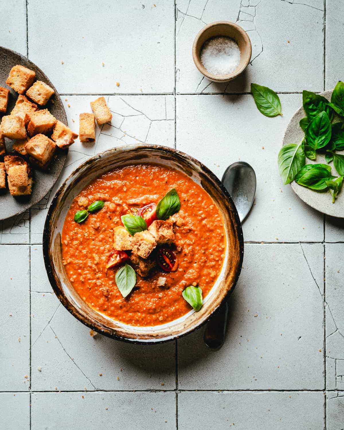 A bowl of tomato soup garnished with croutons and fresh basil leaves. A spoon rests beside the bowl on a tiled surface. Plates with croutons, salt, and more basil are nearby.