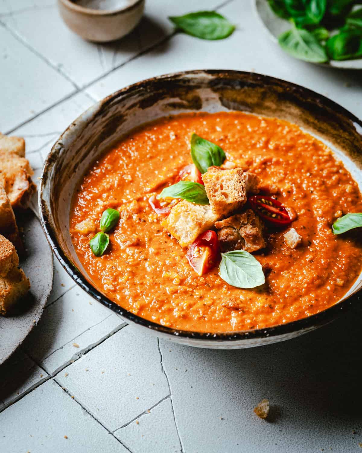 A bowl of creamy tomato soup topped with croutons, red chili slices, and fresh basil leaves. The bowl sits on a textured surface with a beige napkin and additional basil leaves nearby.