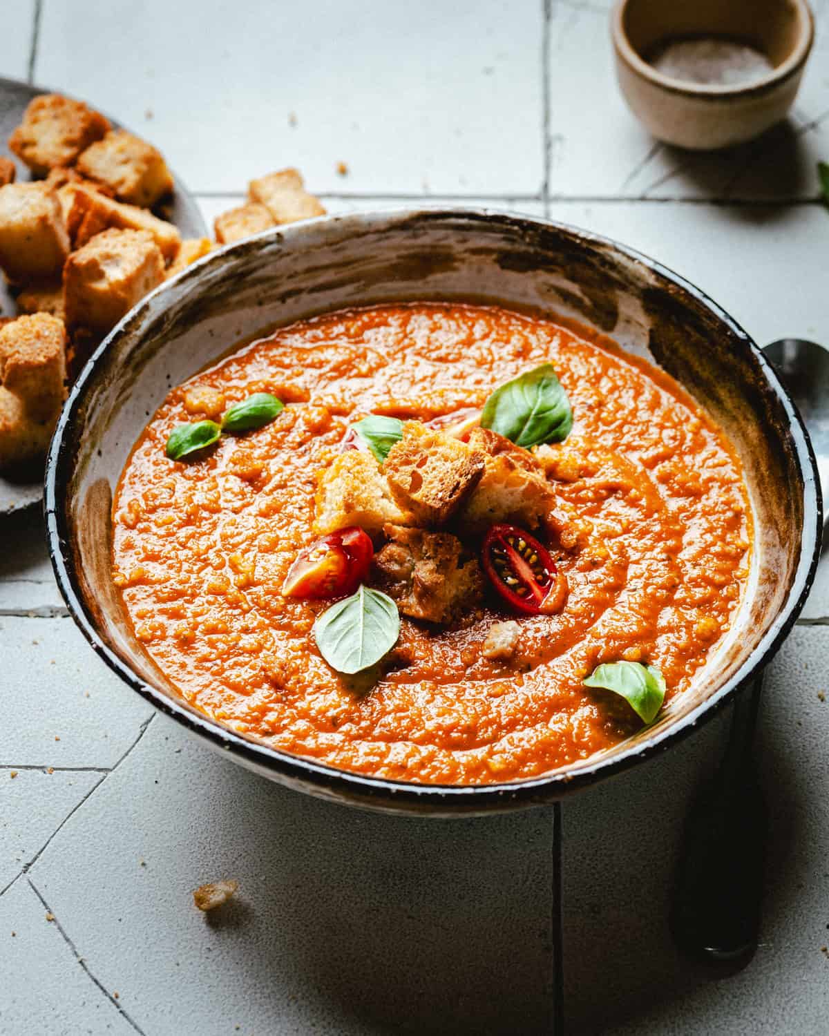 A bowl of creamy tomato soup garnished with croutons, tomato slices, and basil leaves. A small bowl of salt and extra croutons are visible in the background on a tiled surface.