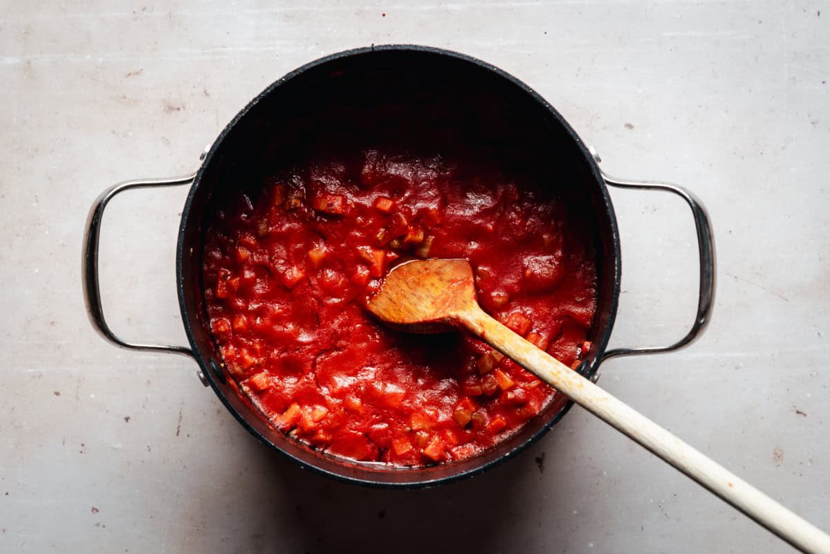 A pot filled with chunky tomato soup on a light-colored surface. A wooden spoon rests inside the pot, stirring the vibrant red sauce.