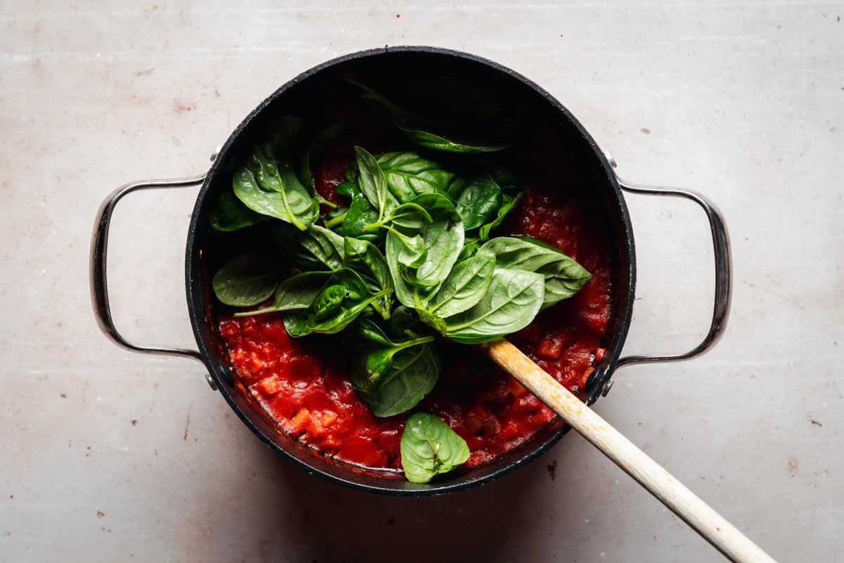 A black pot filled with tomato soup, adorned with fresh basil leaves, sits on a light-colored surface. A wooden spoon rests inside the pot, partially submerged in the sauce.